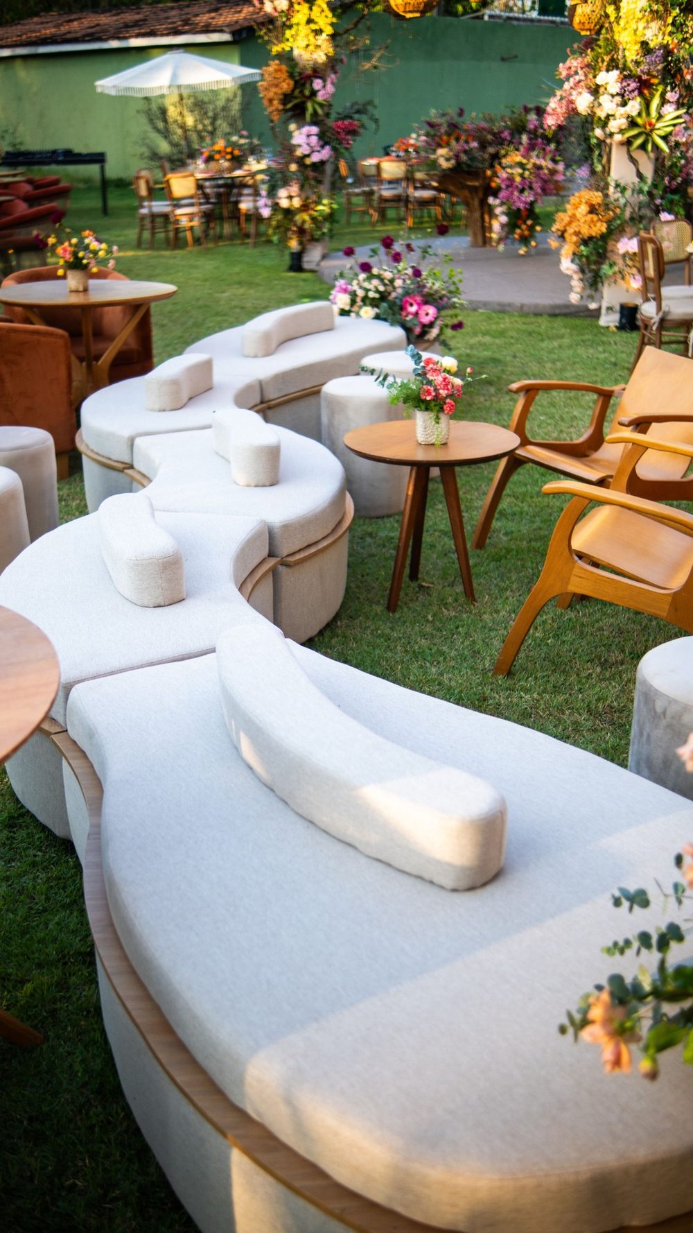 Living curvo tapizado, sillas de madera y mesas auxiliares con flores en un jardín